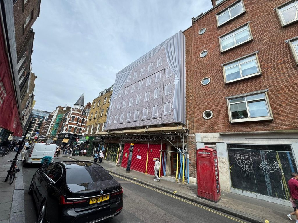 Scaffolding in central London Scaffolding with decorative building wrap covering a property under renovation on a busy London street, with shops, cars, and pedestrians nearby.